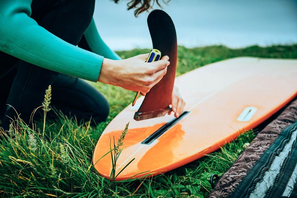 Woman in wetsuit installing a single fin on an orange surfboard in the grass